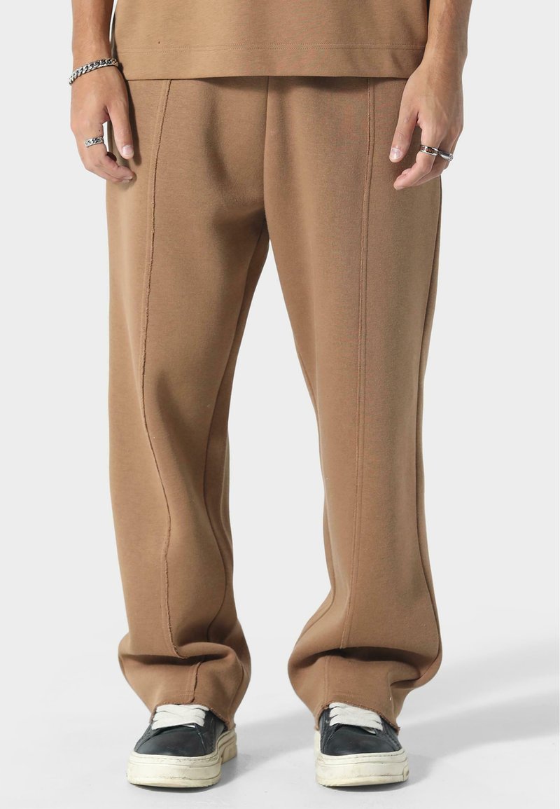 Ruske sweatpants s opuštenim krojem, vidljivim šavovima i glatkom teksturom, upareni s crnim tenisicama i bijelim vezicama.