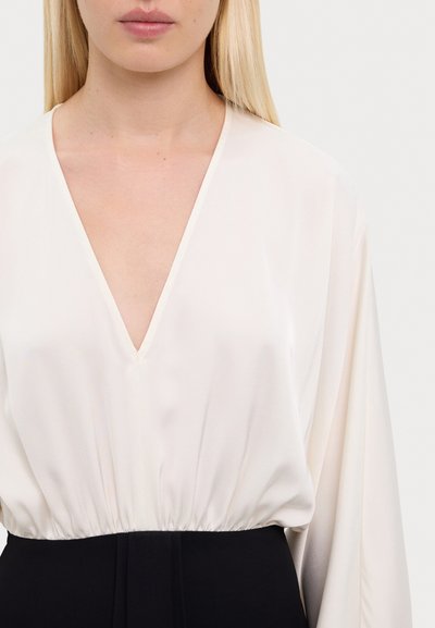 Blouse à col en V en tissu crème doux avec des manches longues et une taille froncée, associée à un bas noir uni. Texture lisse, détails minimaux.