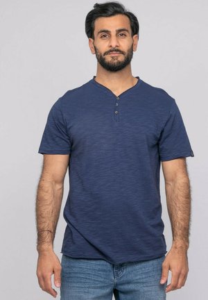 Homme aux cheveux foncés et à la barbe, portant une chemise henley à manches courtes bleue et un jean bleu clair, debout devant un fond gris clair uni.