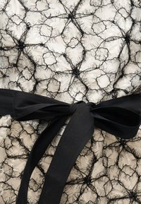 Μαύρη υφασμάτινη δαντέλα με υφή και γεωμετρικά σχέδια πάνω σε ανοιχτό ύφασμα, με διακοσμητική μαύρη υφασμάτινη ζώνη δεμένη.