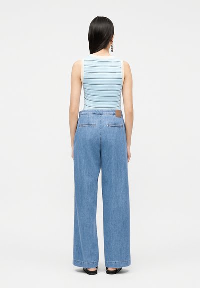 Γυναίκα με μακριά σκούρα μαλλιά, που φοράει αμάνικο λευκό τοπ με μπλε ρίγες και ψηλόμεσα ανοιχτόχρωμα τζιν με φαρδιά μπατζάκια, στέκεται γυρισμένη με την πλάτη.