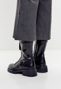 Botine din piele neagră, cu tocuri groase, purtate sub pantaloni gri cu picioare largi, stând pe o suprafață deschisă.