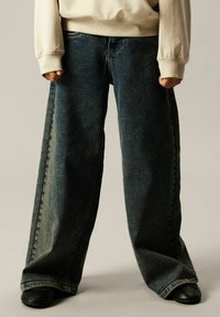 Person iført store mørkeblå jeans og en beige genser som står med knyttede never langs sidene.