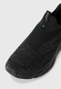 Black slip-on athletic shoe featuring a knitted upper with varying textures, a green accent tab, and a cushioned black sole.