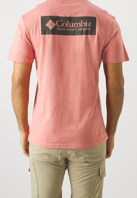 Rosa bomullströja med en svart rektangulär logotyp på baksidan som läser "Columbia Sportswear Company." Kort ärm och rundad halsdesign.