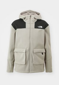 Veste à capuche gris clair avec des panneaux noirs aux épaules, une fermeture éclair devant, deux grandes poches à rabat et le logo The North Face sur la poitrine.
