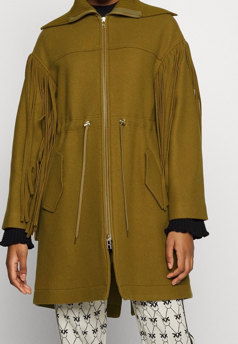Olivgrön kappa med fransdetaljer, dragkedja, sidofickor och en strukturerad krage. Tyget verkar mjukt och texturerat. Mönstrade byxor syns.