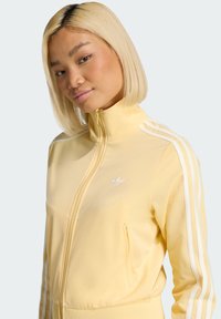 Light yellow athletic jacket with a high collar, white three-stripe detailing on sleeves, and embroidered logo on the chest.