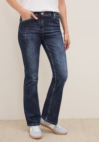 Donkerblauwe denim flared jeans met een middelhoge taille, voorkruiszakken en een knoopsluiting. G gedragen met grijze sneakers en een witte top.
