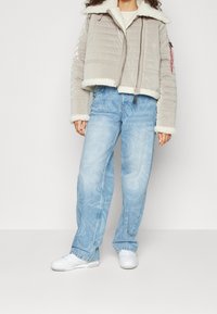 Przycięta beżowa kurtka puchowa z podszyciem z baranka, zestawiona z jasnoniebieskimi, wzorzystymi dżinsami i białymi trampkami. Widoczne zapięcie na zamek i detal z logo.