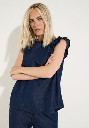 Blonde vrouw met steil haar, gekleed in een donkerblauw mouwloos topje en broek, staand met gekruiste armen tegen een witte achtergrond.
