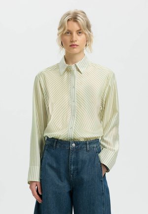 Blonde woman wearing a light green striped long-sleeve shirt tucked into high-waisted dark blue jeans, standing with one hand in pocket.