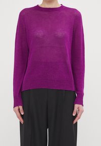 Lila stickad tröja med lång ärm, rund halsringning och ribbad nederkant. Lätt material, något genomskinlig. Svarta byxor synliga nedanför.