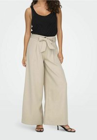 Beige vida byxor med hög midja, försedda med ett knytbälte och en slät tygtextur. Kombinerat med en svart texturerad linne och klackar.