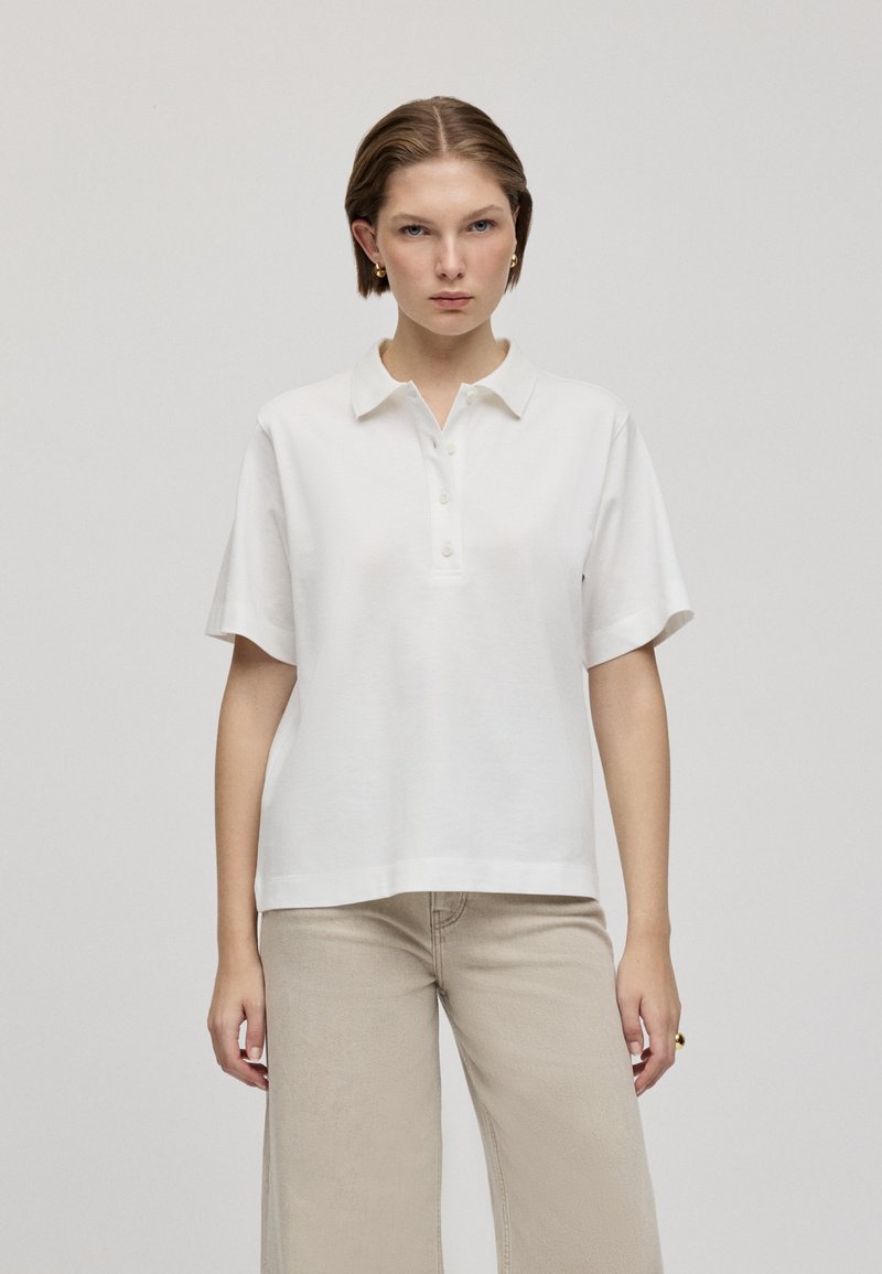 Polo en coton blanc avec col, manches courtes et patte de boutonnage. Présente une coupe classique et une texture douce. Design détaillé et épuré.