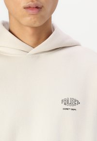 Crème-kleurige hoodie van zachte stof, met een kangoeroezak en een zwart logo met de tekst "PROJECT® CORE™ DEPT." op de borst.