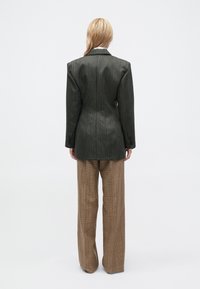 Persoon met blond haar staat met de rug naar ons toe en draagt een donkergrijze streepjesblazer en bruine geruite wijde broek.