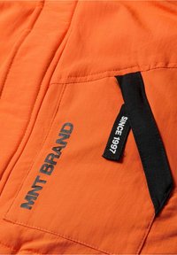 Giacca in tessuto arancione con una tasca con zip nera e dettagli testuali "MNT BRAND" e "SINCE 1997" in colori a contrasto.