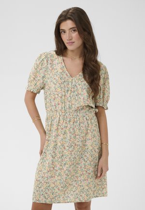 Jonge vrouw met lang bruin haar, gekleed in een lichtgroene bloemenjurk met korte pofmouwen en gouden armbanden, staand tegen een effen achtergrond.