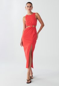 Crvena haljina bez rukava s izrezima sa strane i okruglim detaljem u struku. Ima visok prorez na nozi i mekan, gladak materijal.