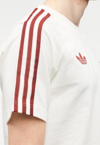 Κοντινό πλάνο ενός ατόμου που φοράει λευκό κοντομάνικο πουκάμισο με τρεις κόκκινες ρίγες στο μανίκι και κόκκινο λογότυπο τριφυλλιού στο στήθος.