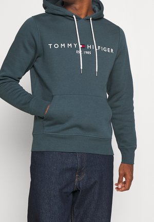 Person trägt einen dunkelgrünen Tommy Hilfiger Hoodie mit weißen Kordeln und Kängurutasche, kombiniert mit dunkelblauen Jeans.