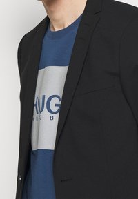 Czarna marynarka o dopasowanym kroju, z pojedynczym zapięciem na guzik i klapami w kształcie notcha, noszona na niebieskiej i szarej koszulce z grafiką, na której widnieje wyraźny tekst.