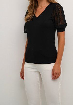 Woman wearing a black V-neck top with lace puff sleeves and white fitted pants, standing against a plain light background.