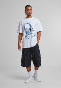 White t-shirt featuring blue headphone graphic with "Miami" text. Black cargo shorts with side pockets and drawstring. White sneakers, crew socks.