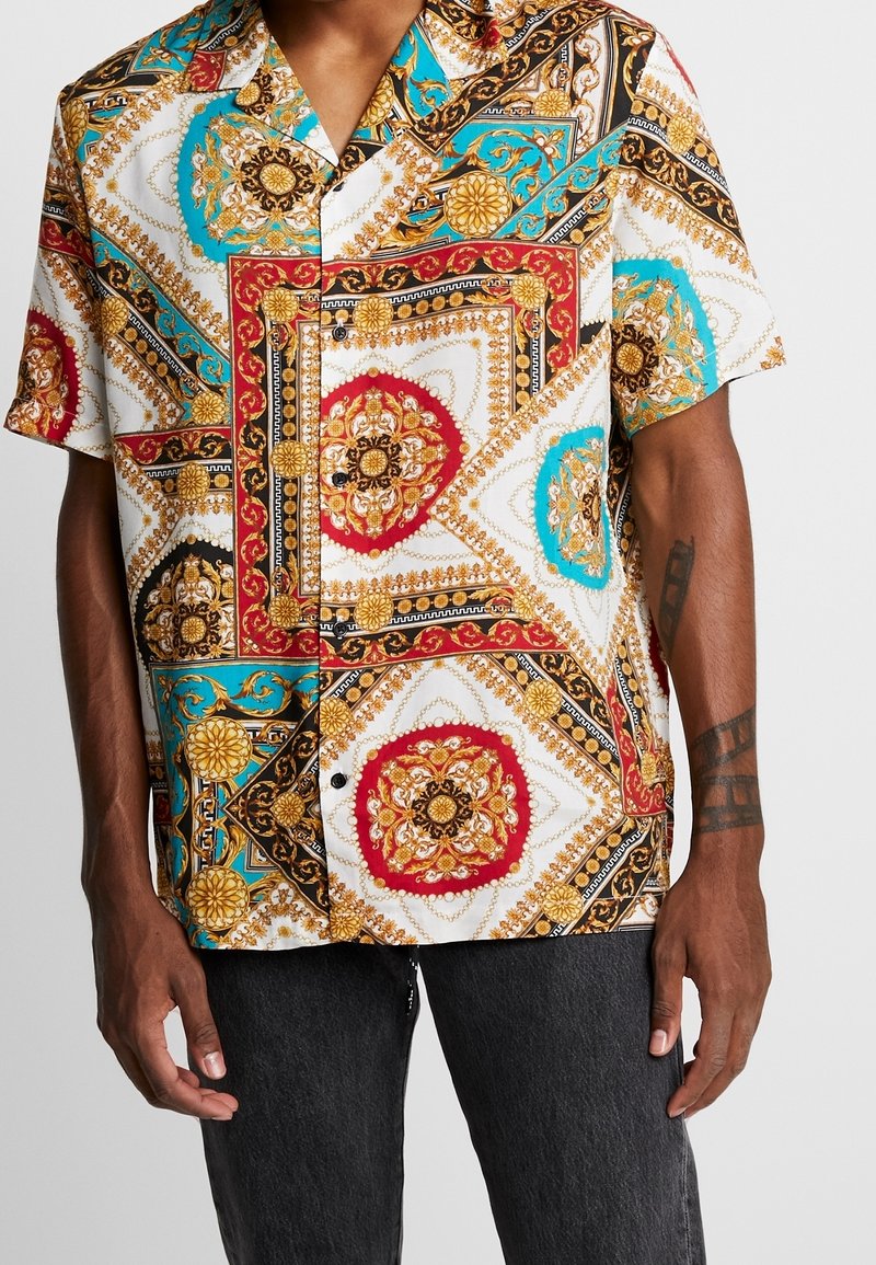 Homme portant une chemise à manches courtes boutonnée avec un imprimé baroque orné rouge, or, noir et turquoise, et un pantalon gris foncé.