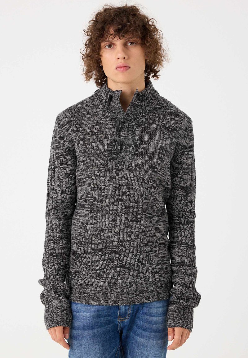 Suéter de punto en negro y gris jaspeado, con un cuello alto, cierre de botón y mangas largas y ajustadas.