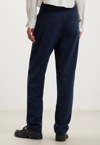Pantaloni navy in tessuto morbido con vestibilità su misura, dotati di due tasche posteriori e un design a gamba dritta e rilassata. Scarpe nere visibili.