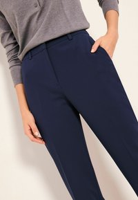 Tmavě modré formální kalhoty z hladké látky, s přizpůsobeným střihem, bočními kapsami a jemnými záhyby vpředu.