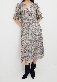 Robe mi-longue fleurie en beige clair avec des motifs bleus, présentant un col en V, des manches à volants, et une silhouette fluide associée à des bottines noires à talons.