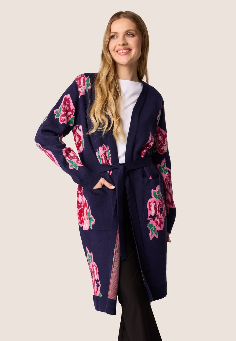 Lange marineblauwe cardigan met bloemenpatroon in roze en groen. Kenmerken zijn een strikceintuur, open voorkant en zijzakken. Zachte gebreide textuur.