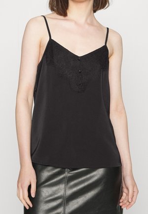 Kobieta w czarnej koszulce na ramiączkach z koronkową lamówką i czarnej skórzanej spódnicy, stojąca na tle jasnego, gładkiego tła.