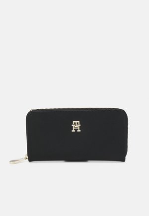 Black wallet with a smooth texture, gold hardware logo on the front, zip closure, and a flat design. Features a wrist strap.