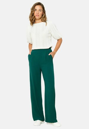 White knitted short-sleeve top with textured pattern paired with green wide-leg trousers. Model stands with hands in pockets, wearing white shoes.