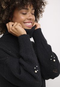 Melnas augstās apkakles džemperis ar ribotu tekstūru, ar pērļu akcentiem uz piedurknēm un brīvu piegriezumu apkakles un ķermeņa daļā.