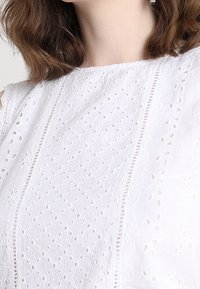 Bílá bavlněná blůza s ozdobnou výšivkou, detaily s výřezem, texturovaným povrchem a vertikálními švy pro strukturu.