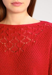 Giovane che indossa un maglione di lana rosso con una texture a pizzo a motivi romboidali sulla parte superiore del petto e sul colletto.