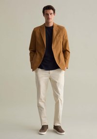 Blazer marrone su una camicia blu navy, pantaloni bianchi e sneaker marroni. Il blazer ha una tasca singola e una vestibilità su misura.