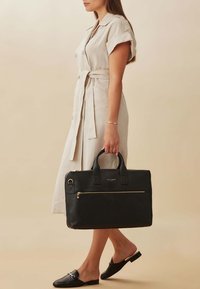 Sac en cuir noir texturé avec deux poignées, une poche avant zippée et des accents dorés. Le sac est rectangulaire avec un design épuré.
