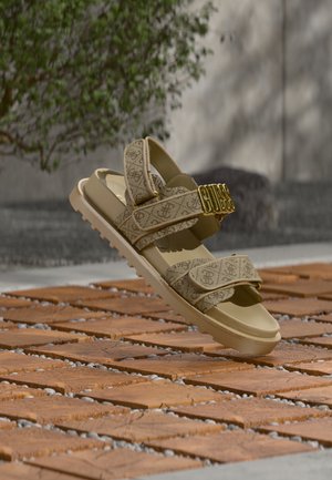 Sandale beige Guess avec brides de marque et boucle logo dorée, présentée en suspension au-dessus de carreaux en terre cuite à motifs en extérieur.