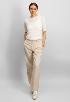 Maglione a maniche corte bianco lavorato a maglia con un motivo testurizzato, abbinato a pantaloni di lino beige. Il modello si trova in posizione casuale con le mani in tasca.