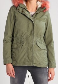 Olivgrüne Parka-Jacke mit pinkem, mit Fell gefüttertem Kapuzenrand, Frontreißverschluss und zwei Brusttaschen. Strukturierter, lässiger Stoff mit verstellbarem Kordelzug.