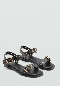 Sandali neri realizzati in materiale sintetico liscio. Presentano accessori in tono oro, fibbie regolabili e accenti decorativi a occhiello. Suola piatta.
