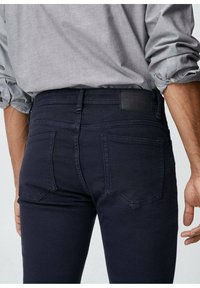 Tummansininen slim fit -farkut, joissa on taskut ja minimaalinen yksityiskohtaisuus. Kangas vaikuttaa pehmeältä, sillä on lievä kiilto ja teksturoitu vyötärö.