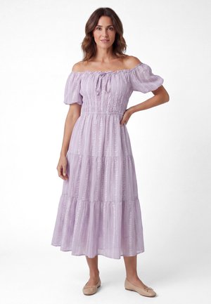 Kvinde står iført en lyse-lilla off-shoulder kjole med puffede ærmer og lagdelt nederdel, kombineret med beige balletsko.