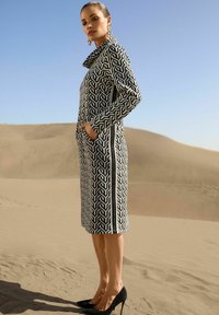Frau in einem langärmeligen Kleid mit schwarz-weißen geometrischen Mustern und hohem Kragen, die in sandigen Dünen der Wüste steht, dazu schwarze High Heels.